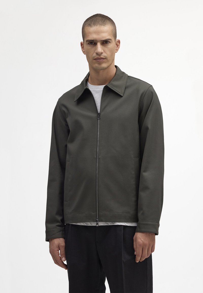 Oliven grøn zip-up jakke med spids krave, letvægtsmateriale og to sidelommer. Båret over en lys grå skjorte, afslappet pasform.