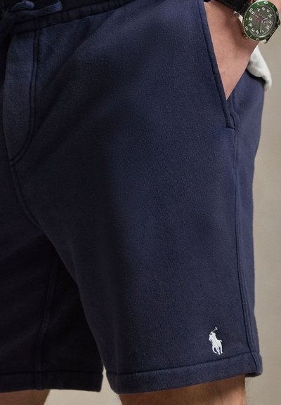 Σκούρα μπλε σορτς με λευκό λογότυπο παίκτη πόλο, χέρι στην τσέπη, φορεμένα με πράσινο και ασημένιο ρολόι χειρός στον αριστερό καρπό.