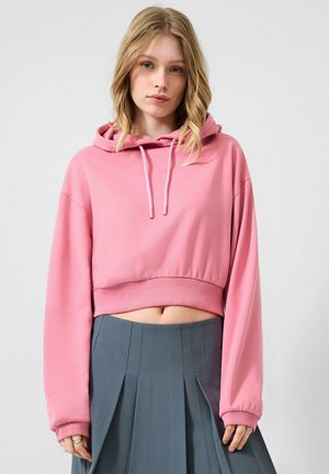 Jeune femme aux cheveux blonds portant un sweat à capuche court rose et une jupe plissée grise taille haute, debout devant un fond clair.