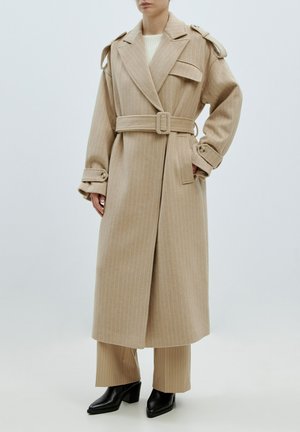 Beige wollen jas met verticale strepen, grote revers, een belted taille en knoopmanchetten. Draagt over bijpassende gestreepte broek en zwarte haklaarzen.