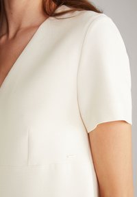 Cremfarbene Bluse mit V-Ausschnitt und kurzen Ärmeln. Sie zeichnet sich durch eine glatte Textur und einen dezenten goldenen Akzent in der Nähe der Naht aus.