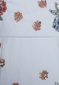 Ljusblått tyg prytt med olika paljetterade blommönster i roséguld, grönt och blått. Texturen verkar slät och mjuk.