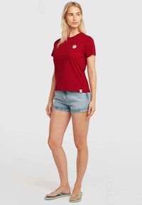 Red short-sleeve t-shirt with a round neckline and a circular logo on the chest, paired with light blue denim shorts and flip-flops.