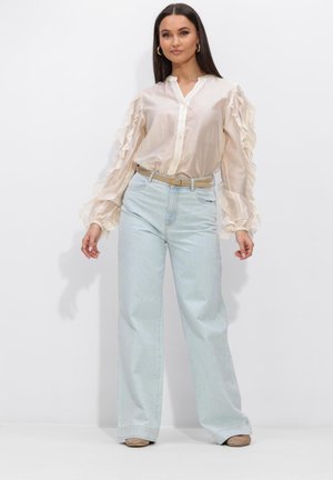 Vrouw staand, gekleed in een doorschijnend beige blouse met geplooide mouwen, lichtblauwe wijd uitlopende spijkerbroek, beige riem en beige schoenen.