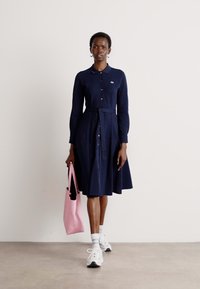 Abito blu navy con colletto, maniche lunghe e cintura annodata, dotato di bottoni lungo la parte anteriore. Abbinate a una borsa tote rosa e scarpe da ginnastica bianche.