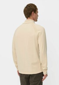 Mann in einem beigen langärmeligen Pullover und dunklen Hosen, der von hinten blickt, mit kurzem braunem Haar vor einem schlichten hellen Hintergrund.