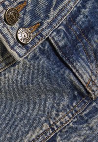 Ύφασμα τζιν σε ανοιχτό μπλε με ξεθωριαστή, φθαρμένη υφή. Περιλαμβάνει μεταλλικά κουμπιά με τη σήμανση «Juicy Couture» και εμφανείς λεπτομέρειες ραφής.