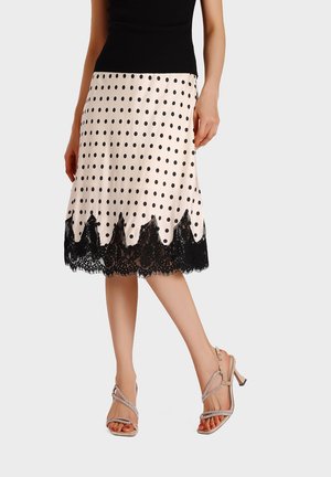 Donna che indossa una gonna color crema a pois con orlo in pizzo nero e sandali beige con tacco alto e cinturini, posata con le gambe incrociate.