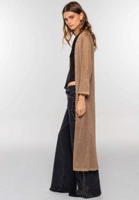 Cárdigan largo de frente abierto en tejido de punto beige con un sutil brillo, combinado con jeans negros de pierna ancha con adornos.