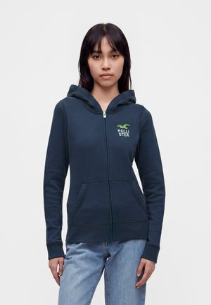 Junge Frau trägt einen marineblauen Zip-Hoodie mit grünem Logo und steht vor einem schlichten hellen Hintergrund.
