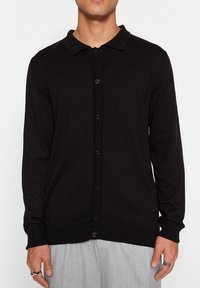 Svart stickad cardigan med krage, knappstängning och ribbade ärmar. Tyget är mjukt och lätt, vilket gör det lämpligt för lager-på-lager.