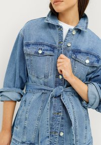Ljusblå denimjacka med midja som kan justeras med bälte, knapprad framtill och två bröstfickor. Tyget har en strukturerad, något blekt look.