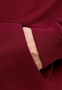 Bordo huppari, jossa on ribattut hihansuut ja tasku, sileä pinta ja näkyvä ompelu yksityiskohtana saumoissa.