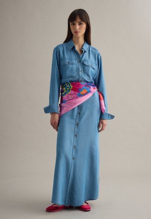 Femme debout portant une longue robe en jean boutonnée, un foulard fleuri rose noué autour de la taille et des chaussures plates roses sur un fond uni.