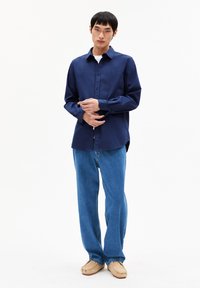 Jonge man staat in een marineblauw overhemd met knopen, blauwe spijkerbroek en beige moccasin schoenen voor een effen witte achtergrond.