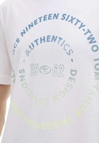 T-shirt en coton blanc arborant un design de texte circulaire multicolore. Les couleurs principales sont le bleu clair, le vert et le jaune, avec des polices et motifs variés.