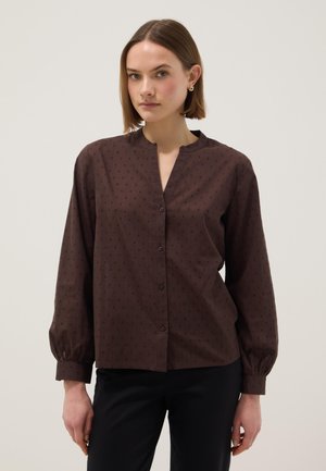 Frau mit kurzen braunen Haaren, die eine dunkelbraune, gemusterte Bluse mit Knöpfen und schwarze Hose trägt, stehend vor einem schlichten Hintergrund.