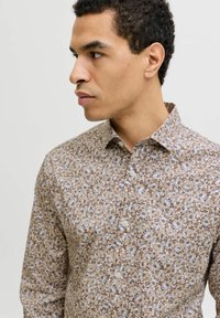 Homme aux cheveux courts et bouclés portant une chemise beige à fleurs boutonnée, regardant vers la gauche sur un fond clair uni.