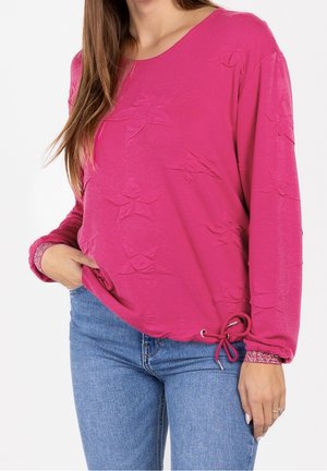 Kvinde iført lyserød langærmet top med teksturerede stjernemønstre og snører i siden, kombineret med blå denimjeans.