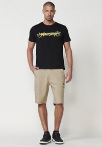 Camiseta de algodón negra con texto gráfico amarillo, combinada con pantalones cortos tipo cargo beige con bolsillos laterales. Zapatillas negras completan el atuendo.