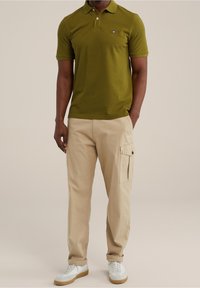 Olivgrön polo med tvåknappsslutning, matchad med krämfärgade cargo-byxor med sidofickor och uppvikta muddar, samt vita sneakers.