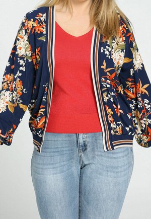 Woman wearing light blue jeans, red V-neck top, and navy blue floral bomber jacket with striped trim.