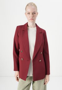 Burgunderfarget dobbeltgjenget blazer i en strukturert passform, med gullknapper og sliwede krager, kombinert med en lysfarget topp.