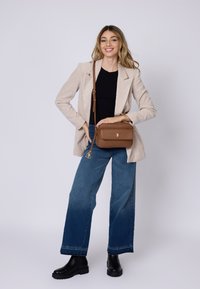 Bolso bandolera de cuero marrón con herrajes dorados, forma rectangular, cierre con cremallera y un charm adjunto. Combinado con una blazer beige y jeans azules.