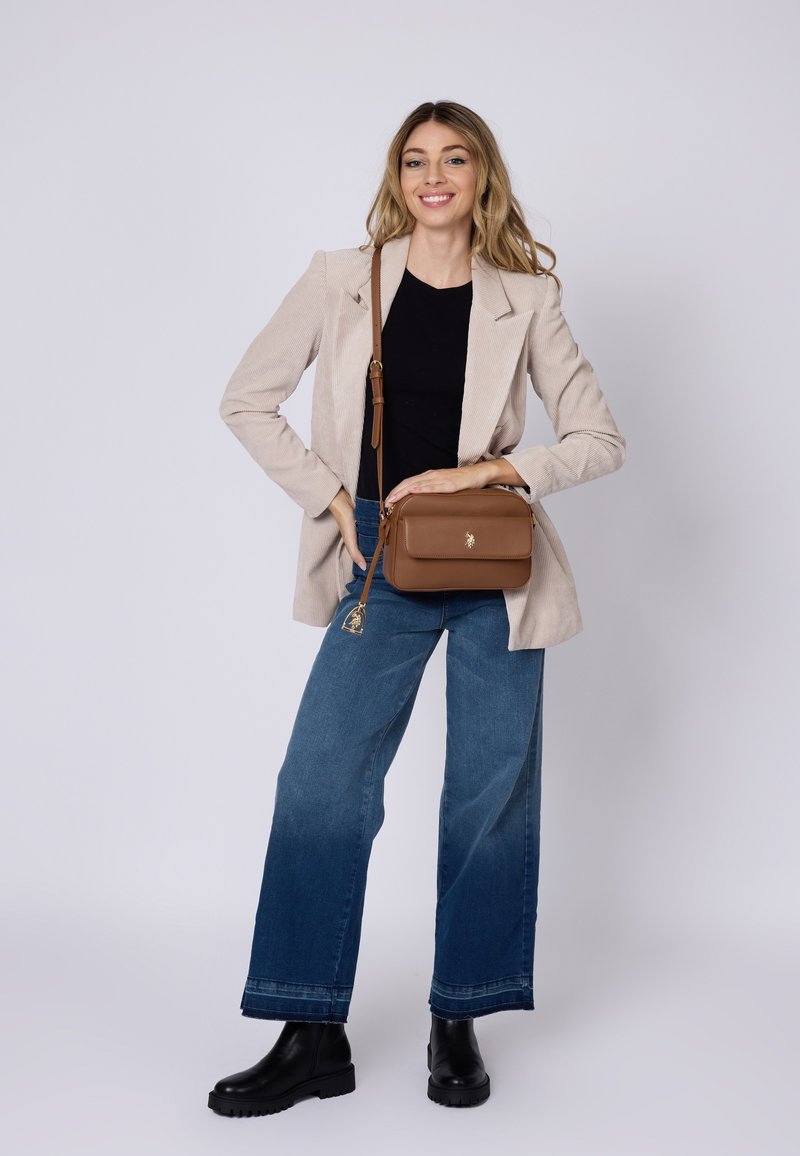 Bolso bandolera de cuero marrón con herrajes dorados, forma rectangular, cierre con cremallera y un charm adjunto. Combinado con una blazer beige y jeans azules.