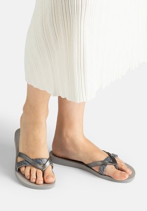 Füße, die grau-schwarze Flip-Flops mit Zebramuster und dicker Sohle tragen, kombiniert mit einem weißen plissierten Rock.