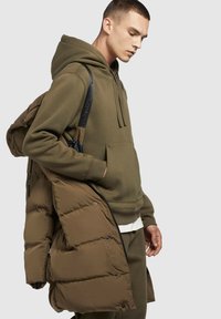 Oliivroheline kapuutsiga särk, millel on ees tasku, on kihitatud pruuni paksu jope alla, millel on nööbid ja sile tekstuur.