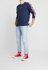 Sweat-shirt navy avec des rayures latérales rouges et blanches, logo brodé, associé à un jean bleu clair et des baskets montantes rouges.