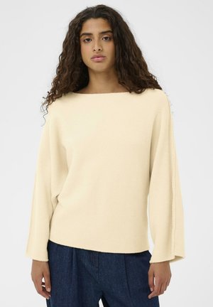 Jeune femme aux longs cheveux bouclés, portant un pull ample couleur crème et un pantalon bleu foncé, debout devant un fond gris uni.