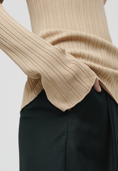 Maglietta beige a maniche lunghe con un fit rilassato, che mette in mostra un tessuto testurizzato. Abbinata a pantaloni scuri su misura, evidenziando il contrasto dei tessuti.