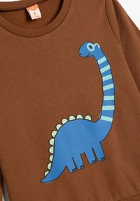 Brązowa bluza z niebieskim motywem dinozaura, z paskowanym karkiem i kolczastym grzbietem, wykonana z miękkiej, teksturowanej tkaniny.