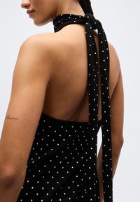 Γυναίκα που φοράει μαύρο φόρεμα με λευκές πουά, χωρίς πλάτη και με το φόρεμα δεμένο με φιόγκο στον λαιμό.