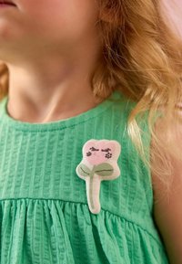 El vestido verde texturizado presenta un broche de tela en forma de flor con un pétalo rosa, una hoja verde y detalles bordados. Diseño simple y juguetón.