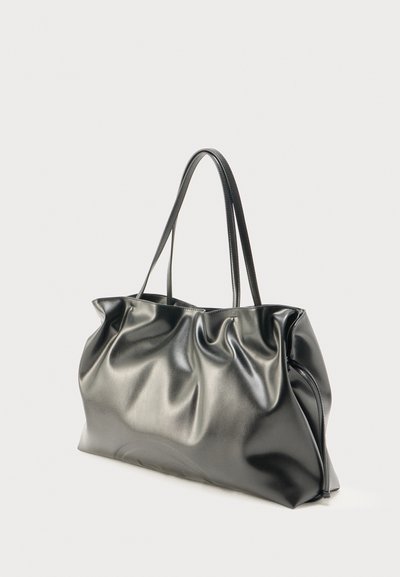 Grand sac cabas en cuir noir avec une texture souple et de longues anses fines doubles sur un fond clair uni.