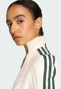 Bijela sportska jakna s zelenim prugastim detaljima na ramenima, visokim ovratnikom i mekanom teksturom. Mali izvezeni logotip na prsima.