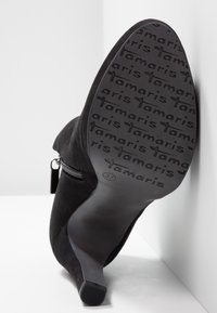 Bottine en daim noir avec un talon effilé, fermeture éclair sur le côté et semelle texturée avec un logo. Taille 37 indiquée sur la semelle.