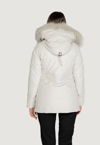 Giacca isolante bianca con cappuccio staccabile foderato in pelliccia sintetica, dettaglio in vita aderente e texture liscia. La vista posteriore mostra una silhouette su misura.