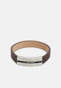Läderarmband i mörkbrunt med en slät yta, med en metallisk silver- och svart logotavla framme.