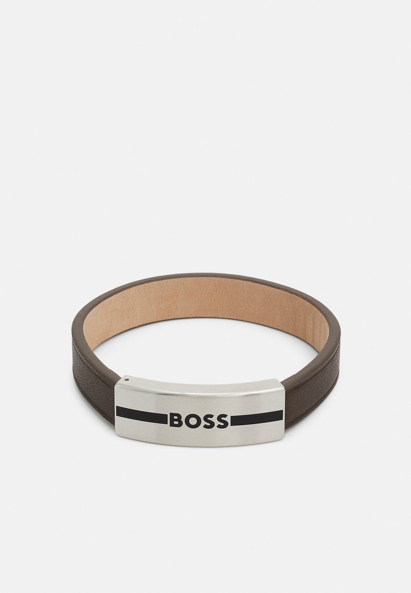 Läderarmband i mörkbrunt med en slät yta, med en metallisk silver- och svart logotavla framme.