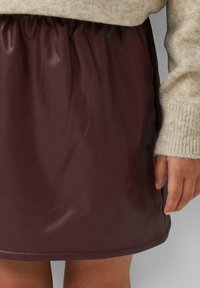 Jupe en simili cuir bordeaux avec une taille froncée. Texture lisse avec un léger éclat. Portée au-dessus du genou, associée à un pull crème.