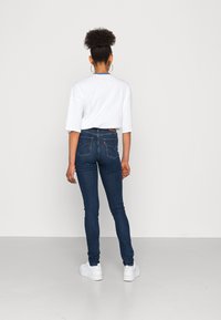 Jeans skinny de ganga escura com uma cintura alta, apresentando bolsos traseiros clássicos e detalhes de costura, combinados com uma blusa branca de manga curta.