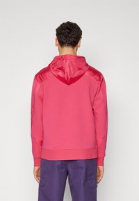 Różowa bluza z kapturem o satynowym wykończeniu na ramionach i kapturze. Posiada prążkowane mankiety i dolny brzeg, charakteryzuje się luźnym krojem i gładką fakturą.