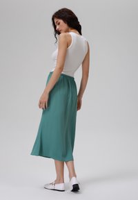 Hvid bomulds tanktop parret med en teal, lang, plisseret nederdel med elastisk talje. Modellen har hvide sko med sorte såler på.