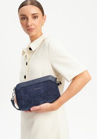Granatowa zamszowa torba na ramię z teksturowaną górą i błyszczącymi akcentami. Posiada zamek błyskawiczny i regulowany pasek.
