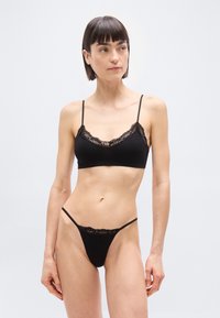 Γυναίκα με κοντά σκουρόχρωμα μαλλιά, φορώντας μαύρο σουτιέν με δαντέλα και εσώρουχο σλιπ που ταιριάζει, στέκεται μπροστά σε απλό φωτεινό φόντο.