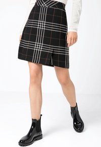 Jupe à carreaux noirs avec des lignes blanches et marron, dotée d'une fente sur le devant. Associée à des bottines noires brillantes et à un ton de peau visible sur les jambes.